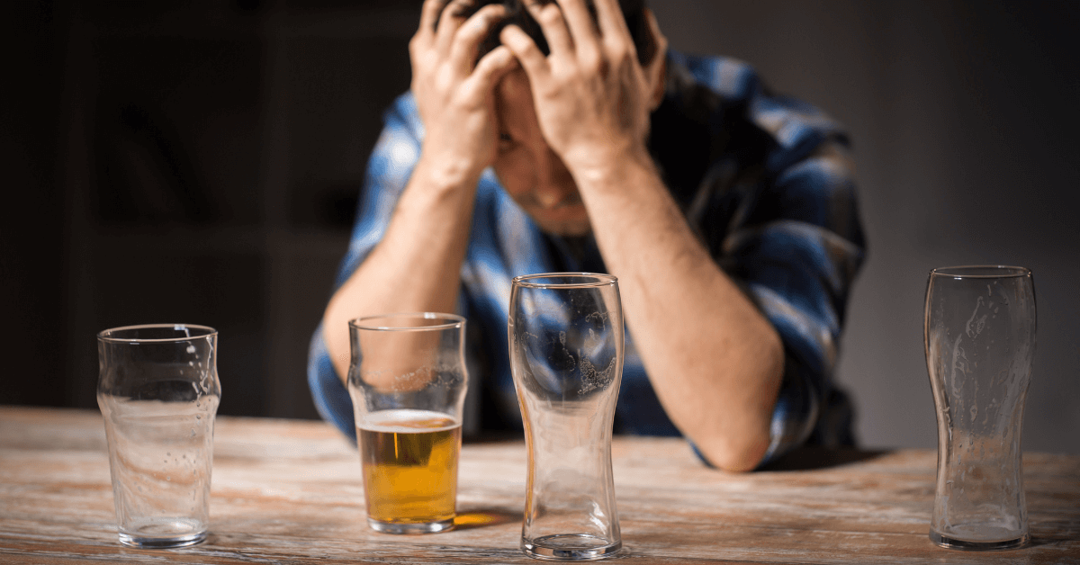 A distressed man in a plaid shirt sits at a wooden table, holding his head in his hands. In front of him are several empty and partially filled beer glasses, suggesting alcohol consumption.