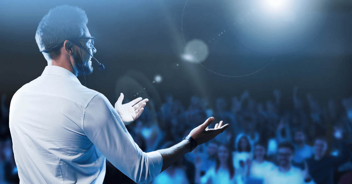 A confident male speaker in a white shirt and headset microphone gestures while addressing a large audience.