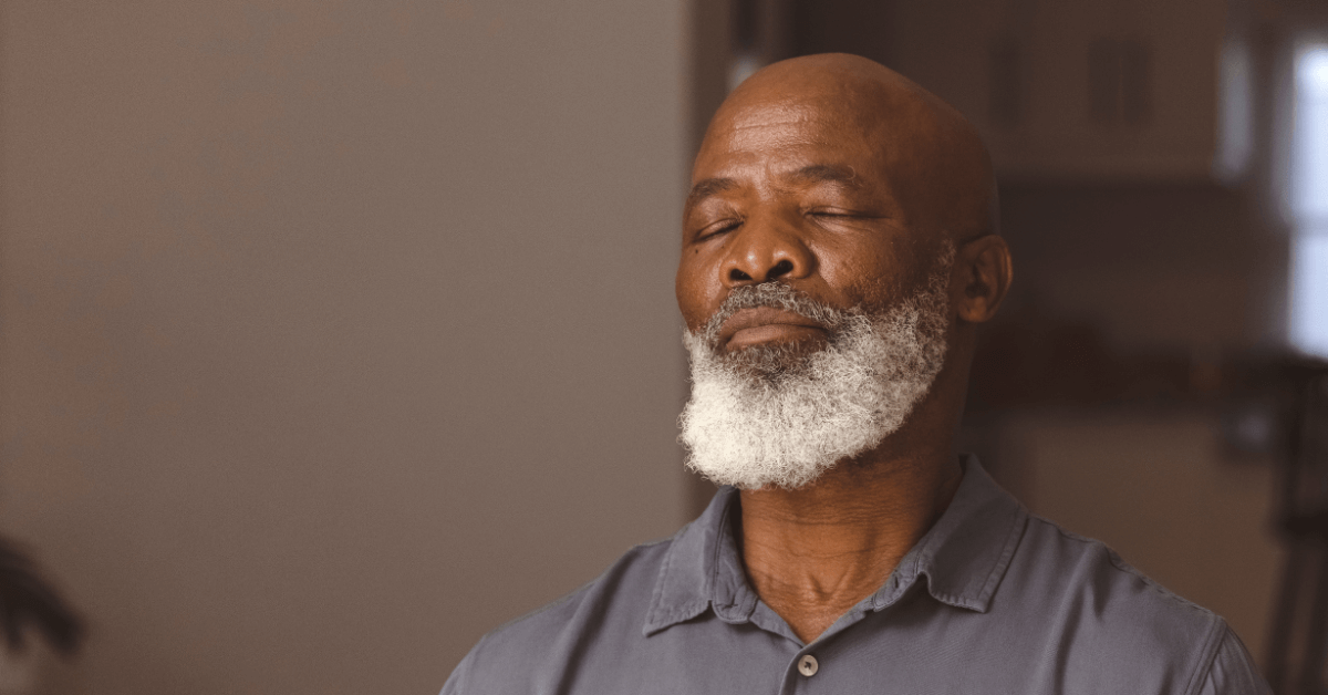 A serene older man with a full white beard and a shaved head sits with his eyes closed, appearing calm and reflective.