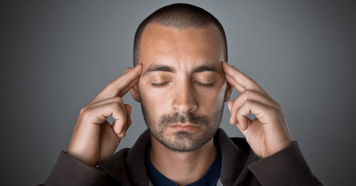 A man with a shaved head and beard, wearing a dark hoodie, closes his eyes and touches his temples with both hands, appearing deep in concentration or meditation against a dark gray background