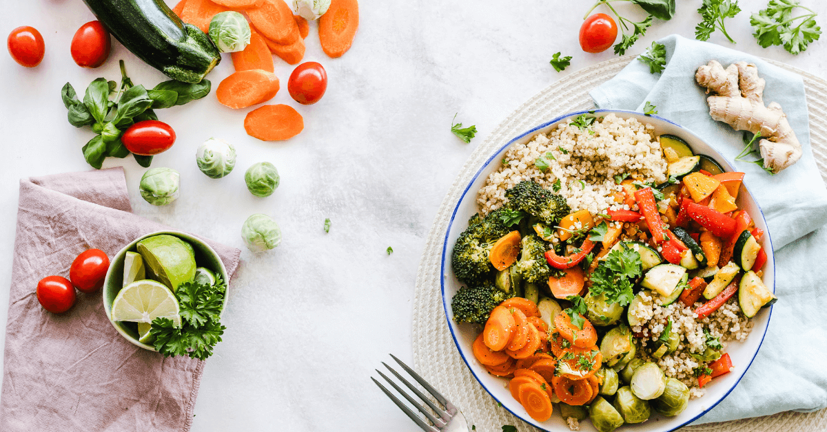 A colourful bowl of quinoa and sautéed vegetables, including broccoli, carrots, zucchini, and bell peppers, surrounded by fresh ingredients like cherry tomatoes, lime, and ginger on a white surface