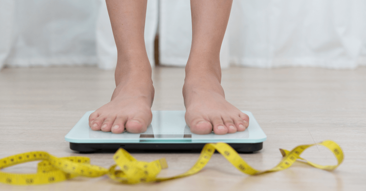 A close-up of a person’s bare feet standing on a digital weighing scale, placed on a light wooden floor. A yellow measuring tape is loosely coiled in the foreground