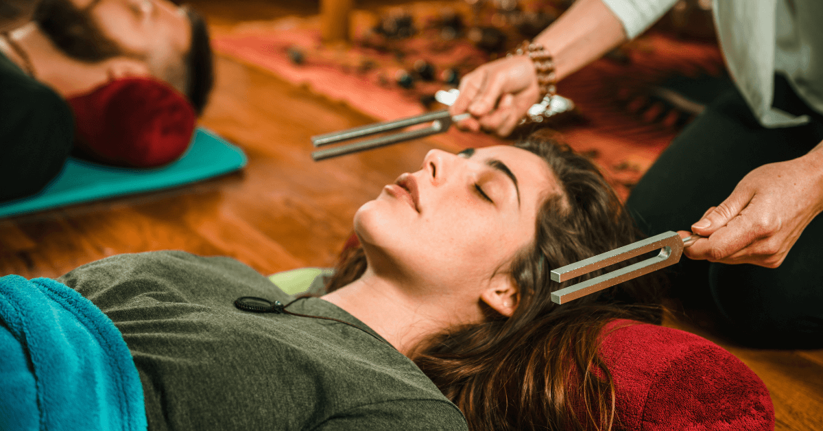 A woman with long brown hair lies on a mat with her eyes closed, appearing deeply relaxed. A practitioner holds two tuning forks near her head, suggesting a hypnotherapy session.